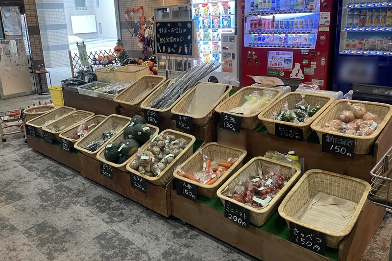 Wicker baskets of bagged vegetables on a stand, each with a small chalkboard price tag in Japanese, with pumpkins and Halloween decorations and drink vending machines behind