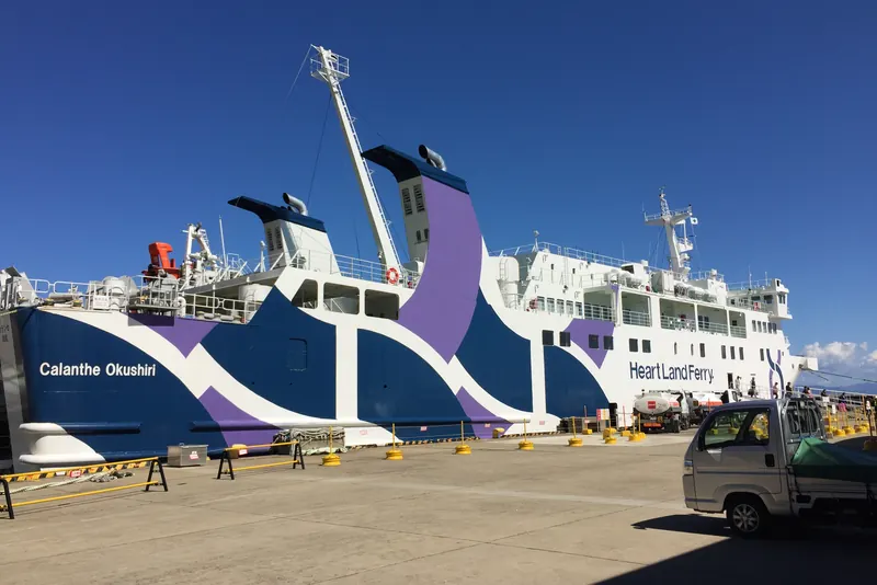 A white ferry with dark blue and purple bands and the name Calanthe Okushiri on its bow moored at a concrete pier, with a small truck parked in the foreground
