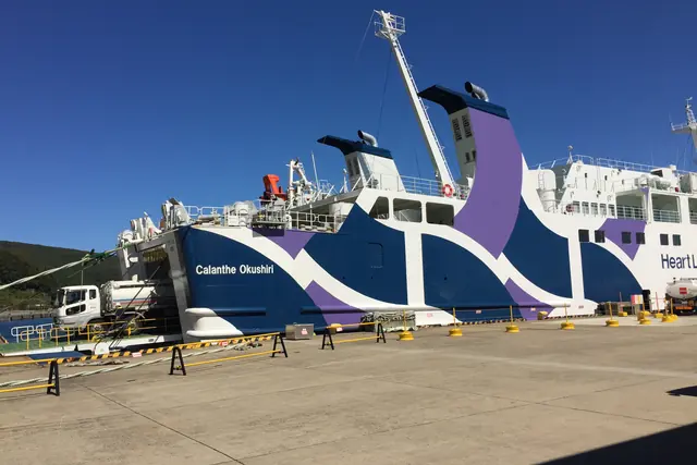 The bow of a white, blue and purple ferry named Calanthe Okushiri at a pier, with a lorry beside its ramp and yellow barriers along the quay