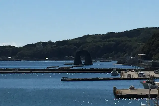 Breakwaters and a few boats in a blue harbour, with a small rock arch standing off a wooded shore behind