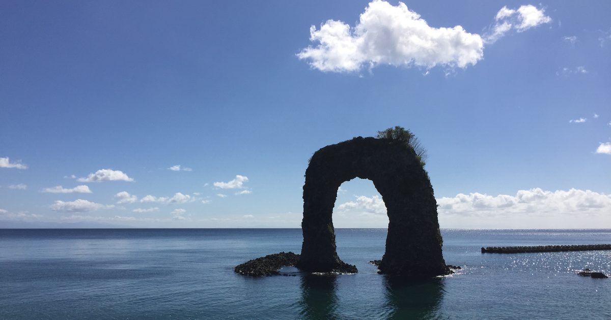 A dark rock arch with shrubs on its top standing in calm sea a short way from shore, with a white cloud above it and a breakwater at the right