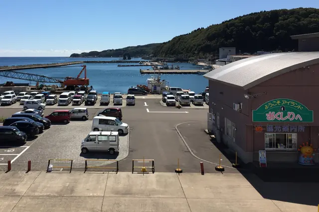 A harbour seen from above with a car park, a building carrying a sign that welcomes visitors to Okushiri and a tourist information office, moored boats, and a small rock arch far along the wooded shore
