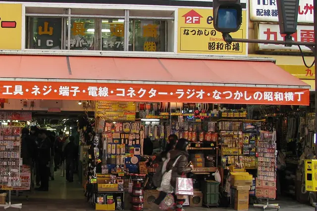 A shop entrance under a red awning with white Japanese lettering, with racks of cables, switches and small electrical parts on both sides and shoppers walking in