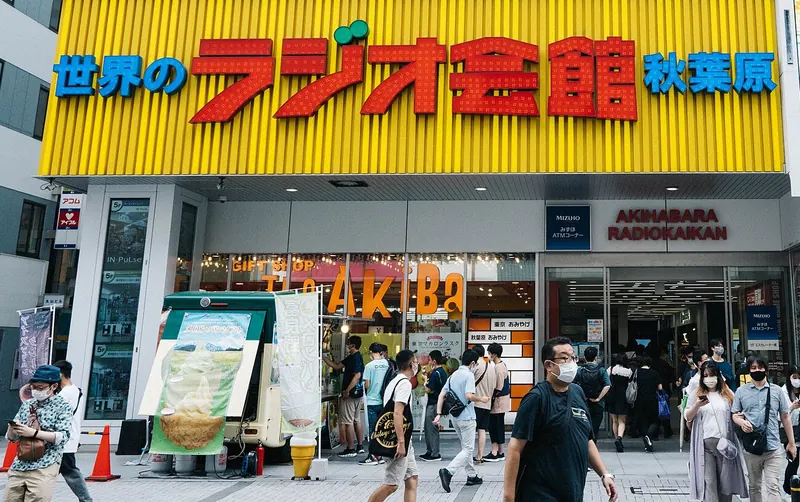 A long yellow sign with red Japanese lettering above the entrance of Akihabara Radio Kaikan, with pedestrians on the pavement below