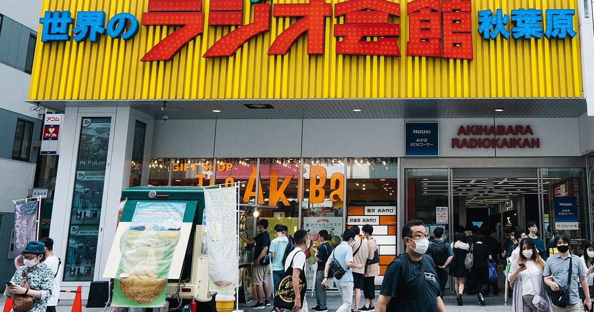 A long yellow sign with red Japanese lettering above the entrance of Akihabara Radio Kaikan, with pedestrians on the pavement below
