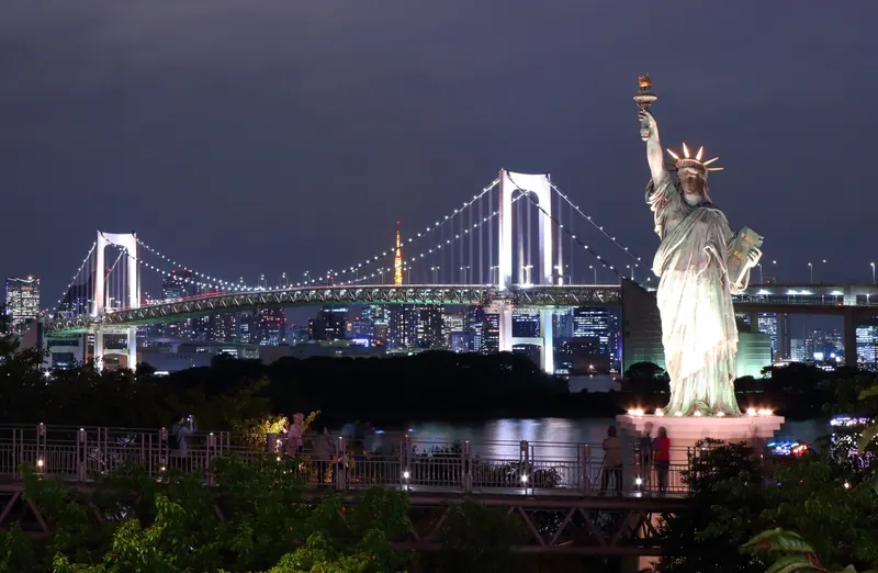 A suspension bridge with white-lit towers at night, a pale green statue holding a torch in front of it and an orange-lit tower in the distance