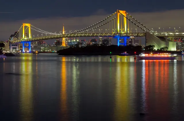 A suspension bridge at night with its towers lit in bands of colour, reflected in the water