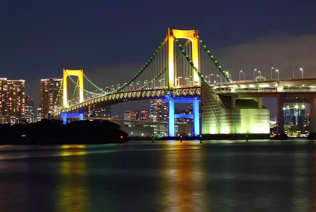 The two towers of a suspension bridge at night, lit yellow above and blue below, with green lights along the cables and reflections on the water