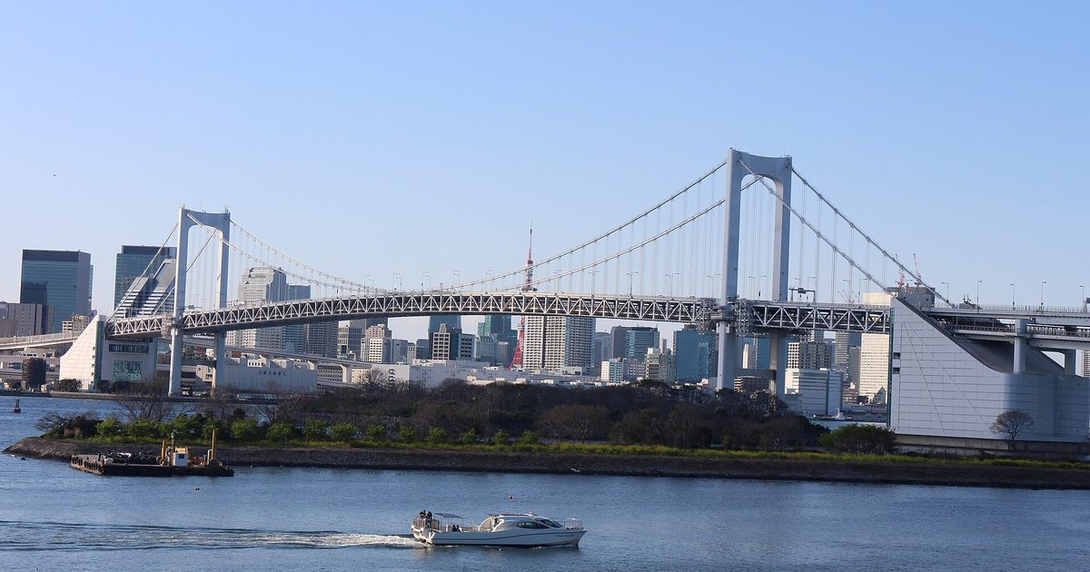 A white suspension bridge with two towers crossing a bay on a clear day, with a red-and-white tower and tall buildings behind it