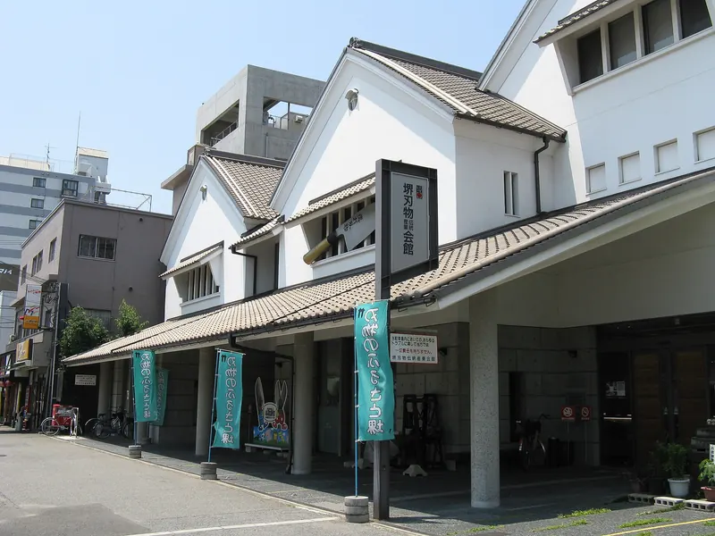 A white-walled building with a tiled roof and banners by its entrance, on a street in Sakai