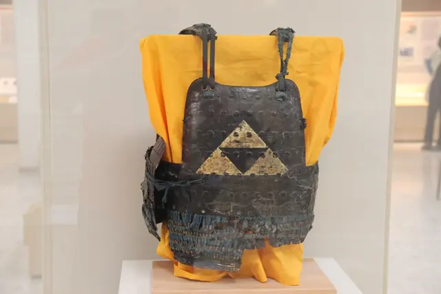 An iron cuirass with a gold crest over yellow cloth in a museum case