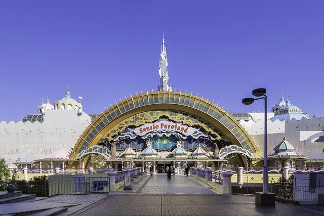 The entrance of Sanrio Puroland under a large rainbow-coloured arch with a Sanrio Puroland sign, a white spire behind it and a wide walkway with a few visitors leading up to the ticket windows