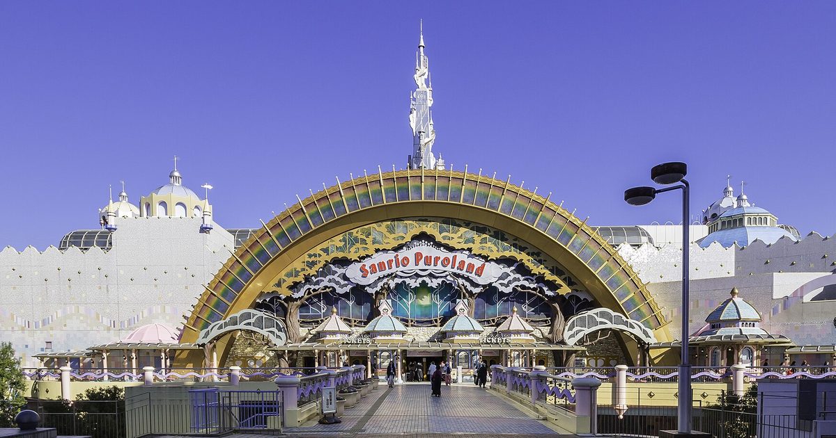 The entrance of Sanrio Puroland under a large rainbow-coloured arch with a Sanrio Puroland sign, a white spire behind it and a wide walkway with a few visitors leading up to the ticket windows