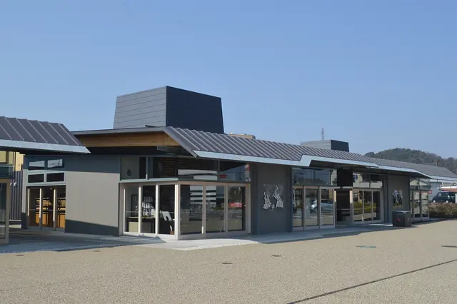 A low modern building with large windows and a grey roof behind a wide paved forecourt