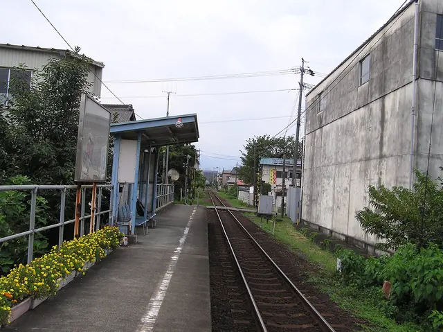 A single-track station platform with a small shelter and yellow flowers along its edge
