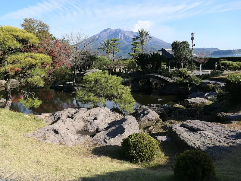 A garden of rocks, a pond, pines and palms with the volcano Sakurajima rising across the bay