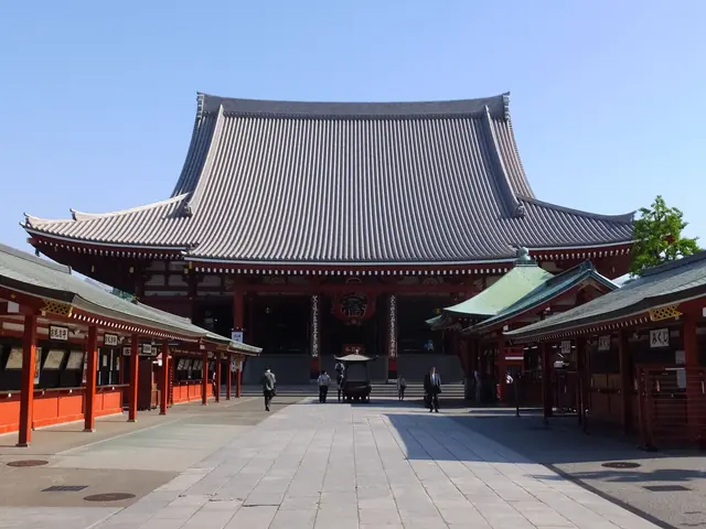 Photo: Tak1701d (CC BY-SA 3.0) The main hall of Senso-ji with a huge grey tiled roof and red pillars across a paved court under a blue sky
