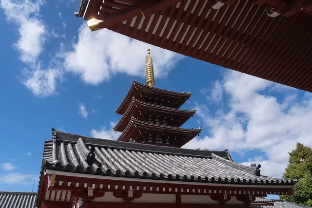 A five-storey pagoda seen from below, framed by a red-raftered roof against a blue sky with white clouds