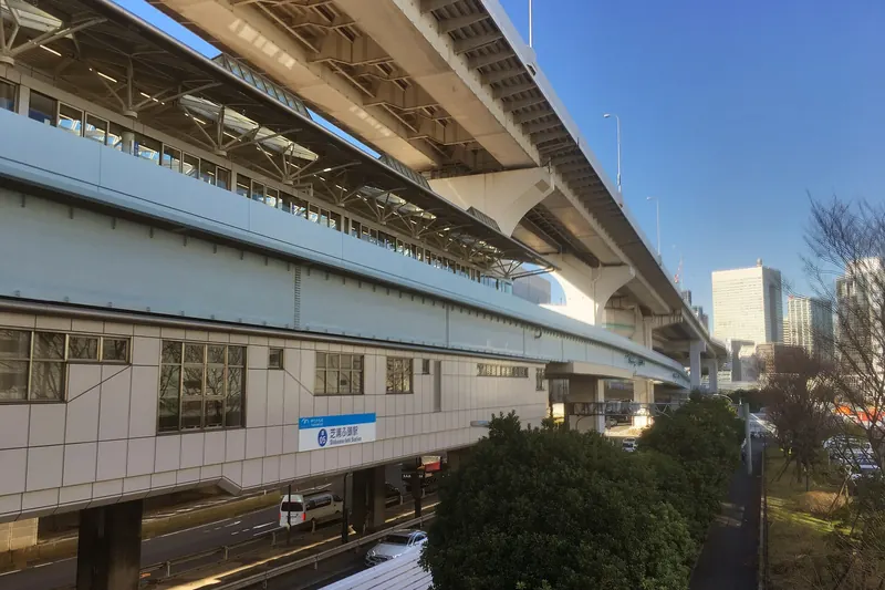 An elevated station building with a blue sign, with trees in front and office towers behind