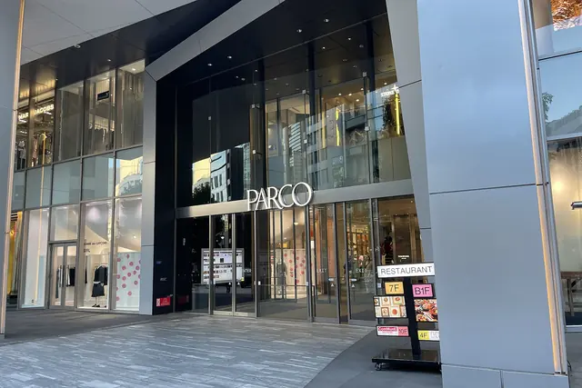 The glass street entrance of a tall modern building with the name PARCO in white letters above the doors and a restaurant floor guide standing beside a silver column