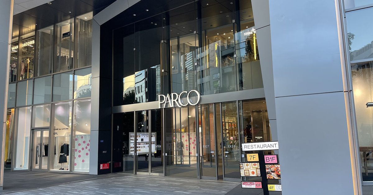 The glass street entrance of a tall modern building with the name PARCO in white letters above the doors and a restaurant floor guide standing beside a silver column