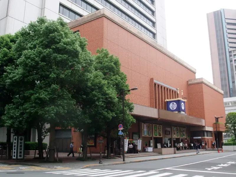 The brown brick-faced Shinbashi Enbujo theatre on a street corner, with large trees in front and a navy crest sign on the wall