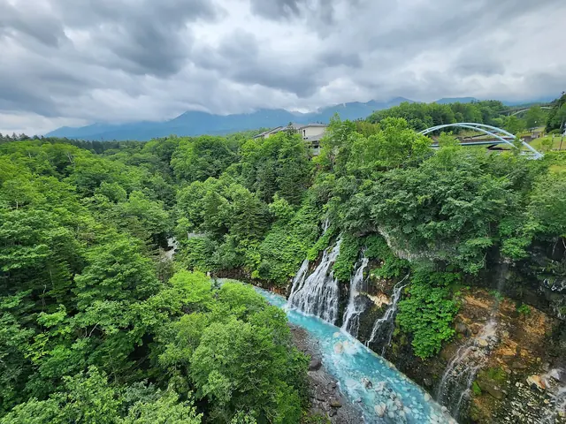 Falls dropping into a wooded river gorge crossed by an arch bridge, with mountains under cloud behind