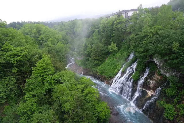 Wide view of a green gorge with water falling down the rock face into the river and a building on the rim above