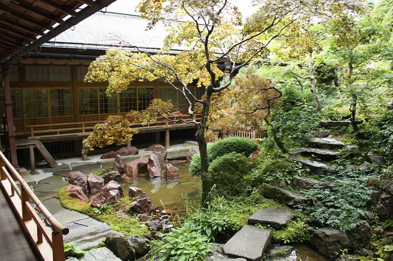 A small garden of rocks, a pond, maples and stone steps seen from a wooden veranda