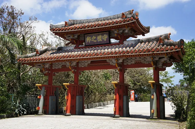 A two-tier wooden gate with red-tiled roofs and red pillars, with trees behind and a blue sky