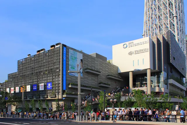 A low grey shopping complex with a Tokyo Solamachi sign at the foot of a white lattice tower, with a long line of people on the pavement in front