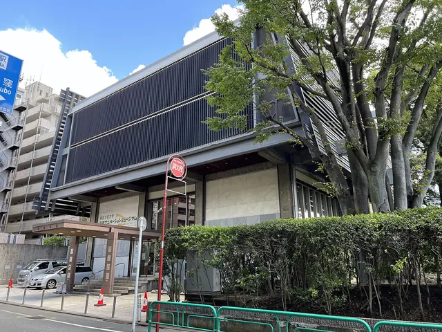 A building with a wall of black vertical slats beside a large tree and a hedge, seen from the road