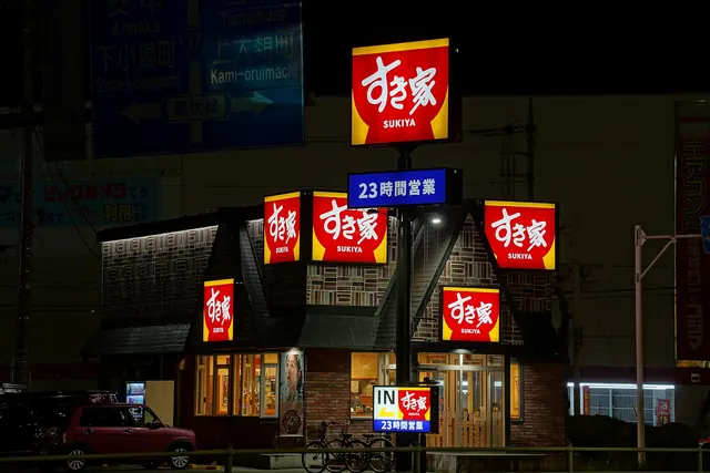 A Sukiya restaurant lit up at night with red and white signs on the roof