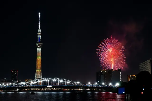 A red firework burst beside the lit Tokyo Skytree above an arched bridge at night