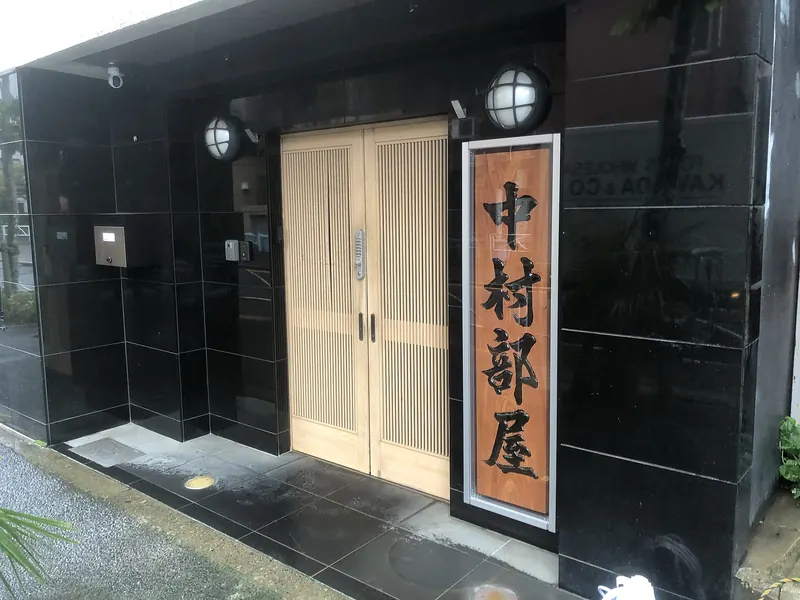 A pale wooden sliding door set in a black tiled wall, beside a tall wooden board carved with the stable name in black characters