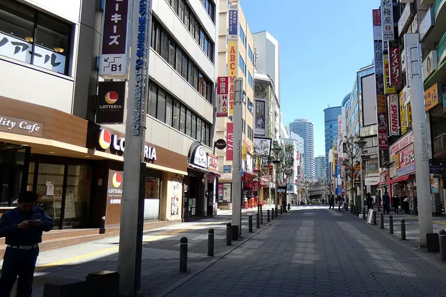 A wide paved pedestrian street with bollards and tall shop signs on both sides, leading towards glass towers in the distance, with almost nobody on it