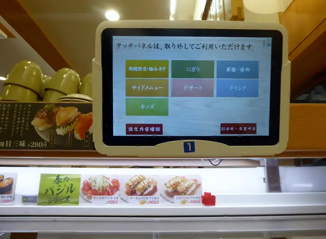 A touch-screen tablet above a conveyor belt showing menu categories, with menu photos below