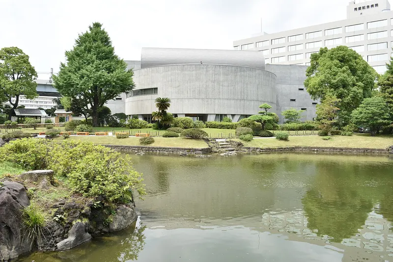 A curved pale grey museum building behind a garden pond and trees