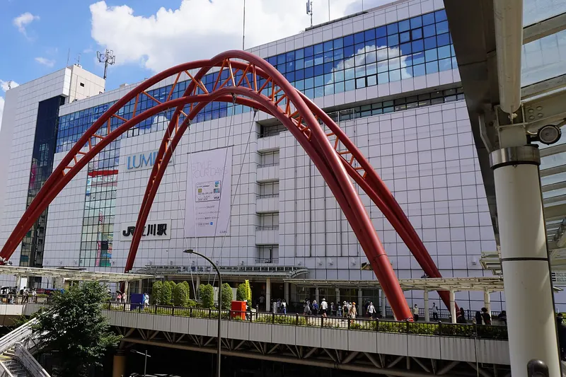 A tall red arch standing over a raised pedestrian deck in front of a white station building, with the station name in Japanese on the wall