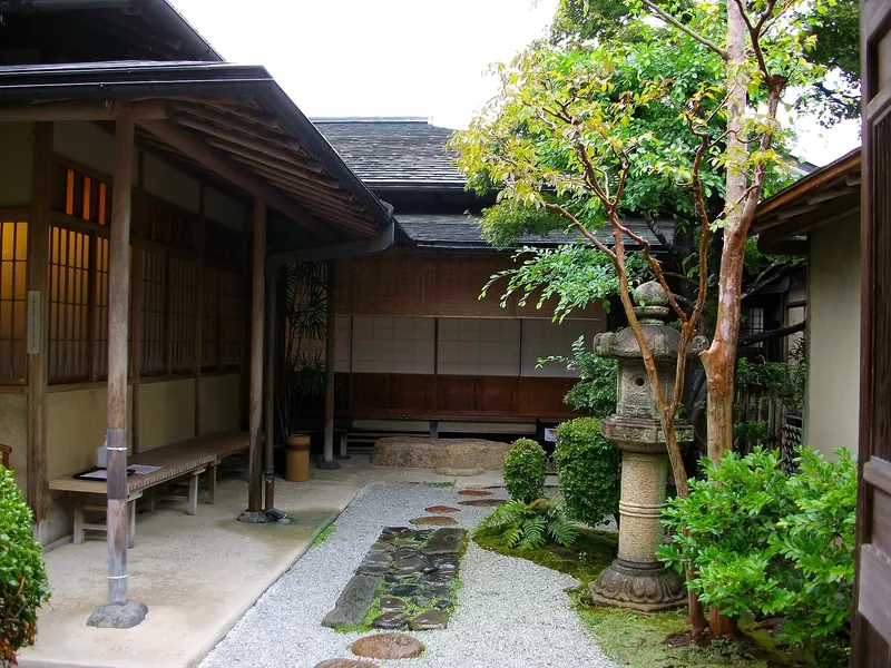 The wooden veranda of a tea house beside a gravel path with stepping stones, a stone lantern and green trees