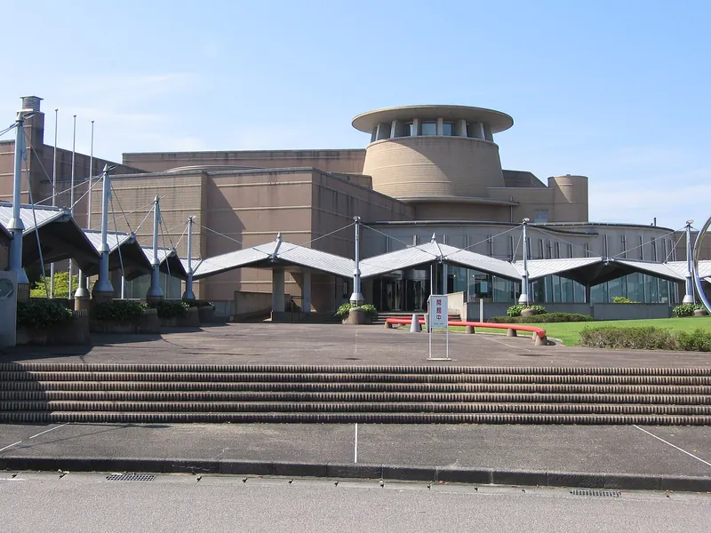 The Takaoka Art Museum, a low brown building with a round tower and a row of white canopies above wide front steps