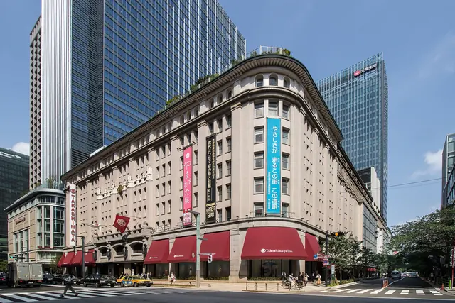 A stone department store with a rounded corner, red awnings along the street and long vertical banners on its front, with glass towers rising behind it under a blue sky