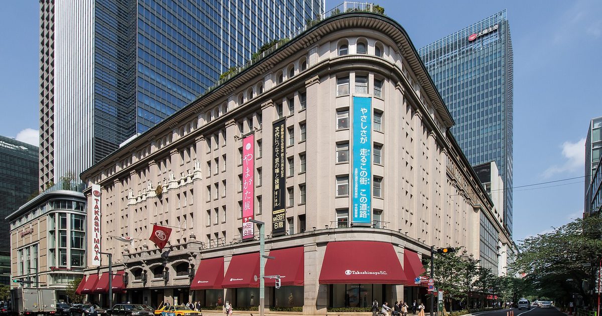 A stone department store with a rounded corner, red awnings along the street and long vertical banners on its front, with glass towers rising behind it under a blue sky