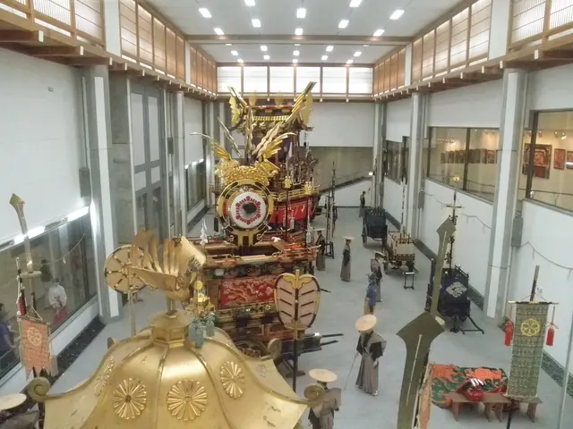 An ornate festival float with a gold-crested roof in a long exhibition hall, with the gilded roof of a portable shrine in the foreground
