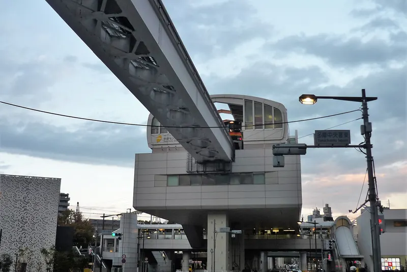 The concrete Tama Center station of the Tama Toshi Monorail raised above a road junction, with the monorail track running into it and evening sky behind