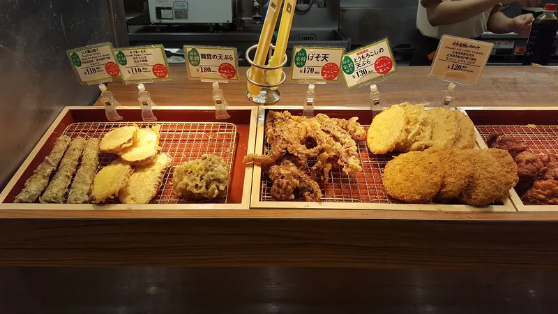 Trays of tempura on wire racks behind a counter with small price cards and tongs