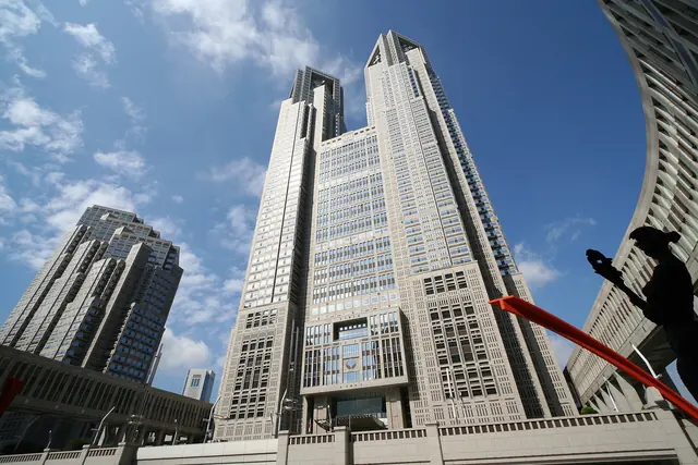 The twin towers of the Tokyo Metropolitan Government Building seen from below against a blue sky with clouds