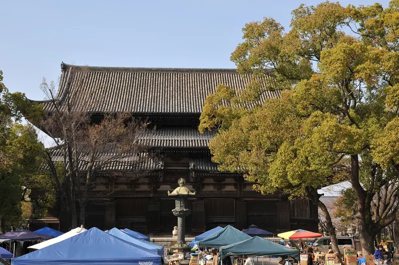 A large wooden temple hall with a tiled roof behind trees, with blue and green market tents in front