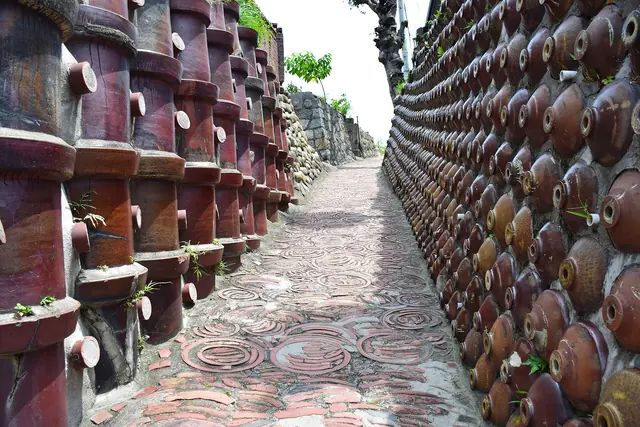 A narrow lane paved with round tiles, running between walls of stacked brown clay pipes and rows of round brown jars