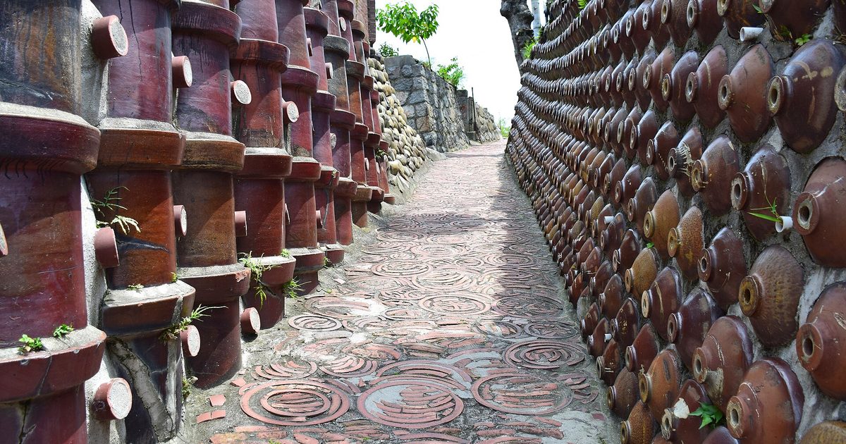 A narrow lane paved with round tiles, running between walls of stacked brown clay pipes and rows of round brown jars